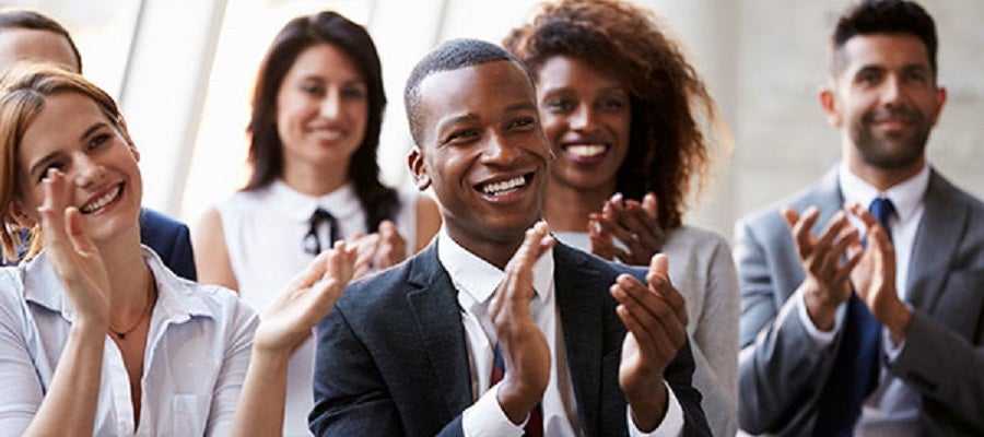 A diverse group of businesspeople siting in three rows smiling and clapping.