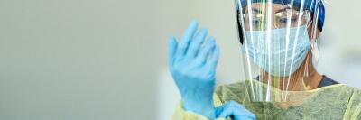 Project Firstline homepage carousel banner. A female clinician wear a gown, surgical mask, and face shield dons a pair of nitrile gloves.